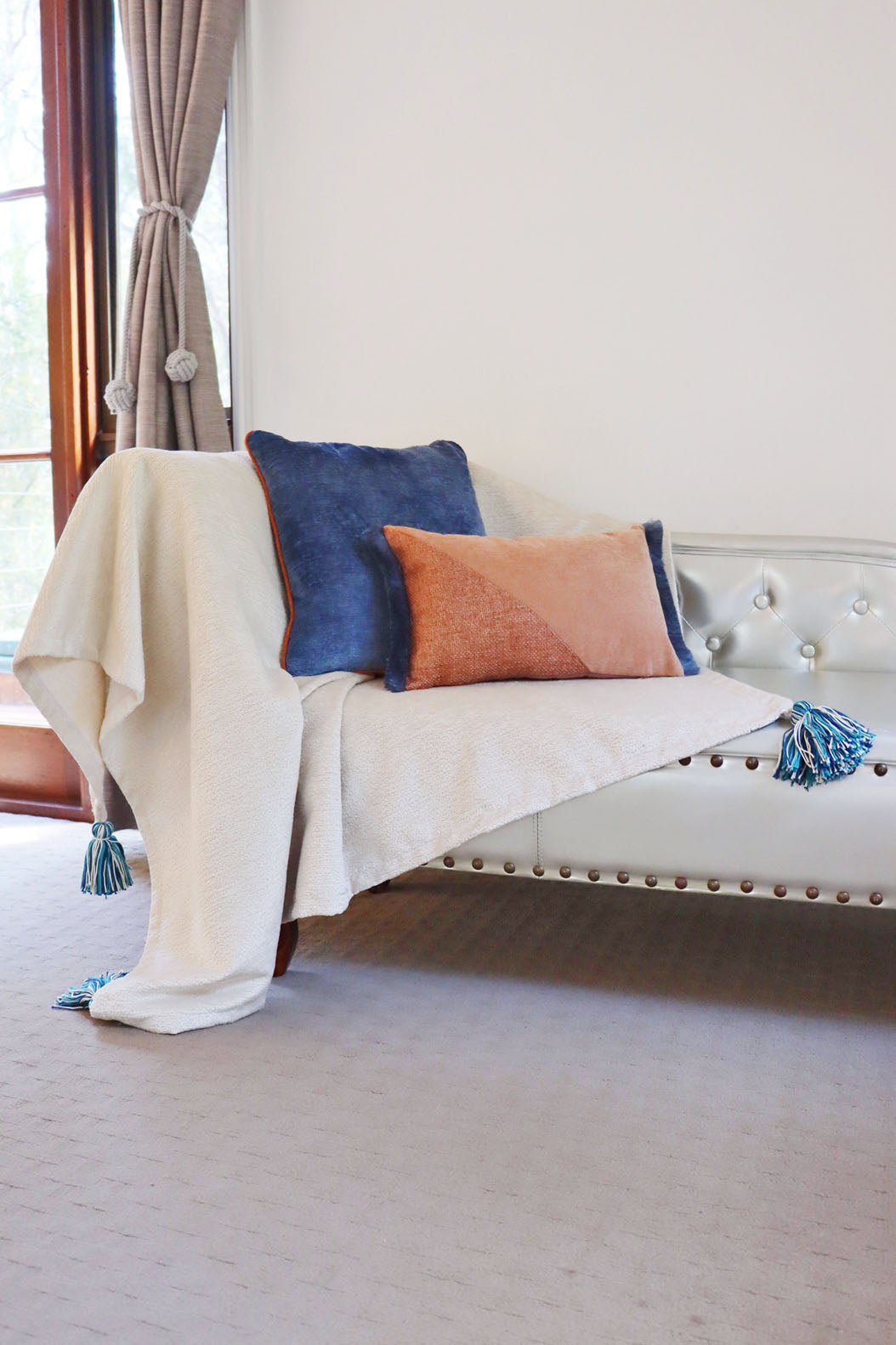 White sofa with a white throw blanket and colorful chenille cushion cover with contrasting piping in a room with a window and curtains.