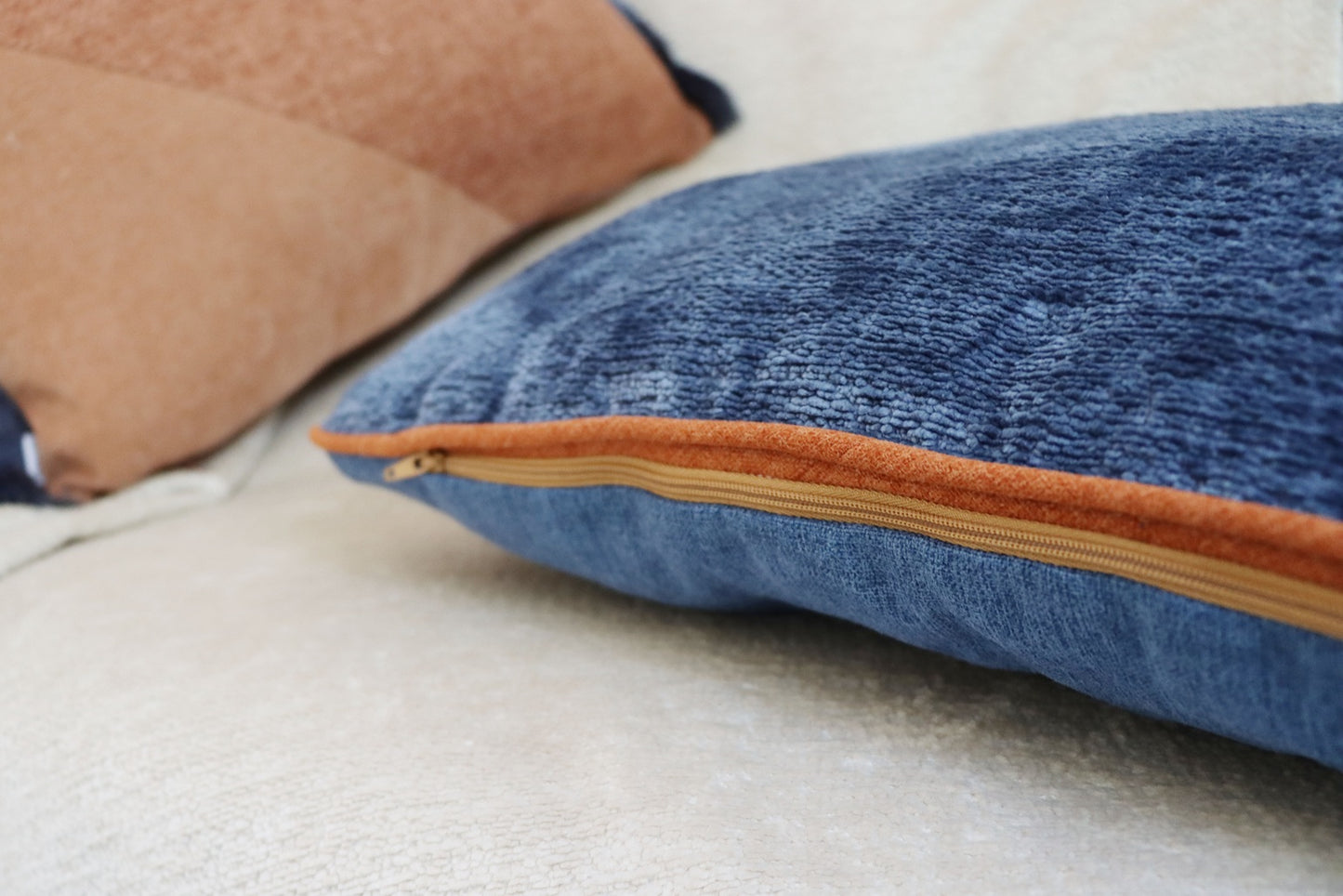Close-up of a blue chenille cushion cover with contrasting piping with a brown zipper on a beige surface.