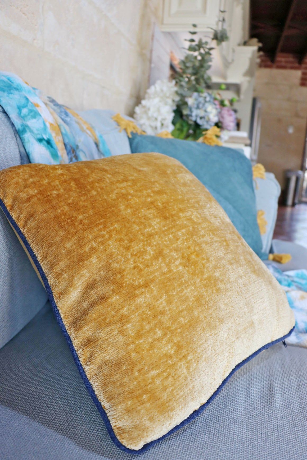Gold chenille cushion cover with contrasting piping on a couch with a blurred indoor background
