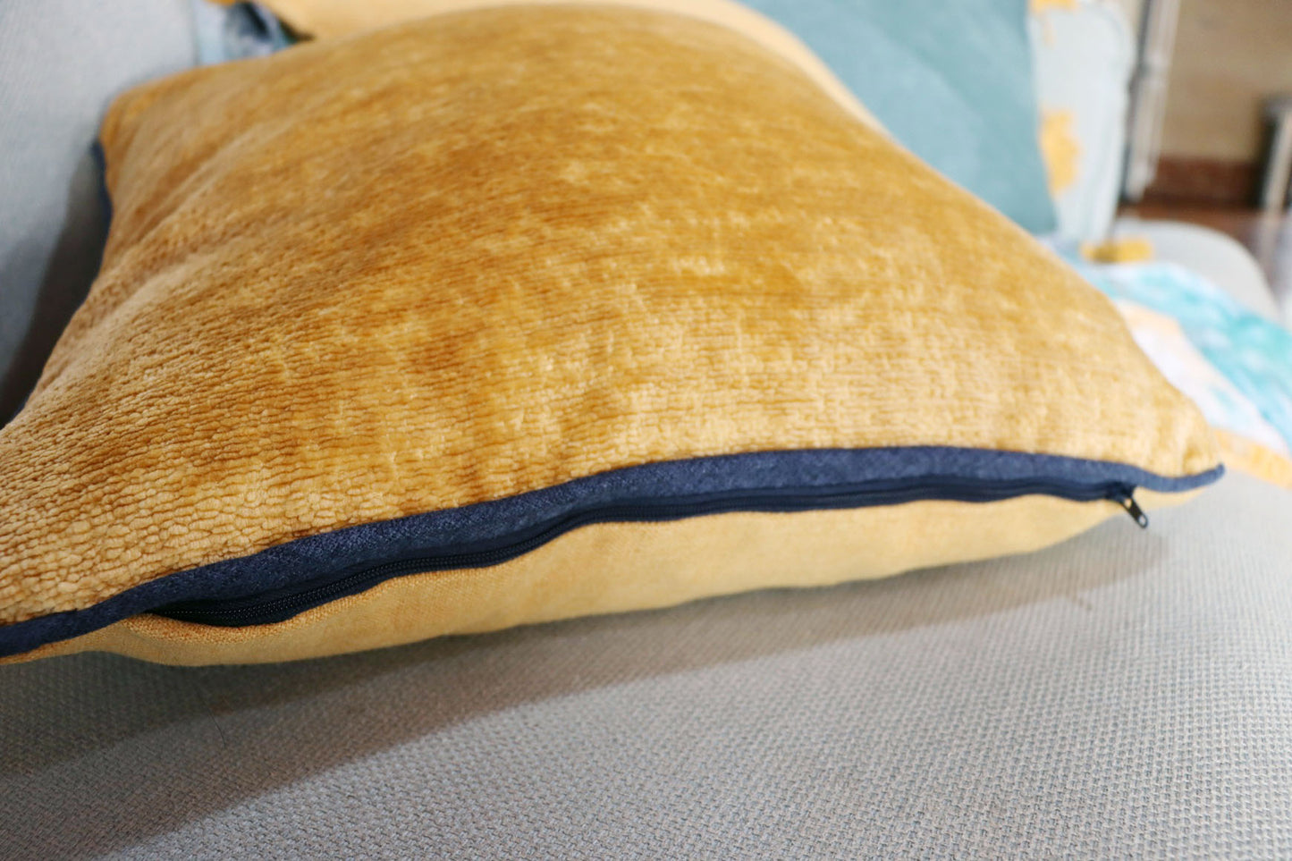 Gold chenille cushion cover with contrasting piping on a gray surface
