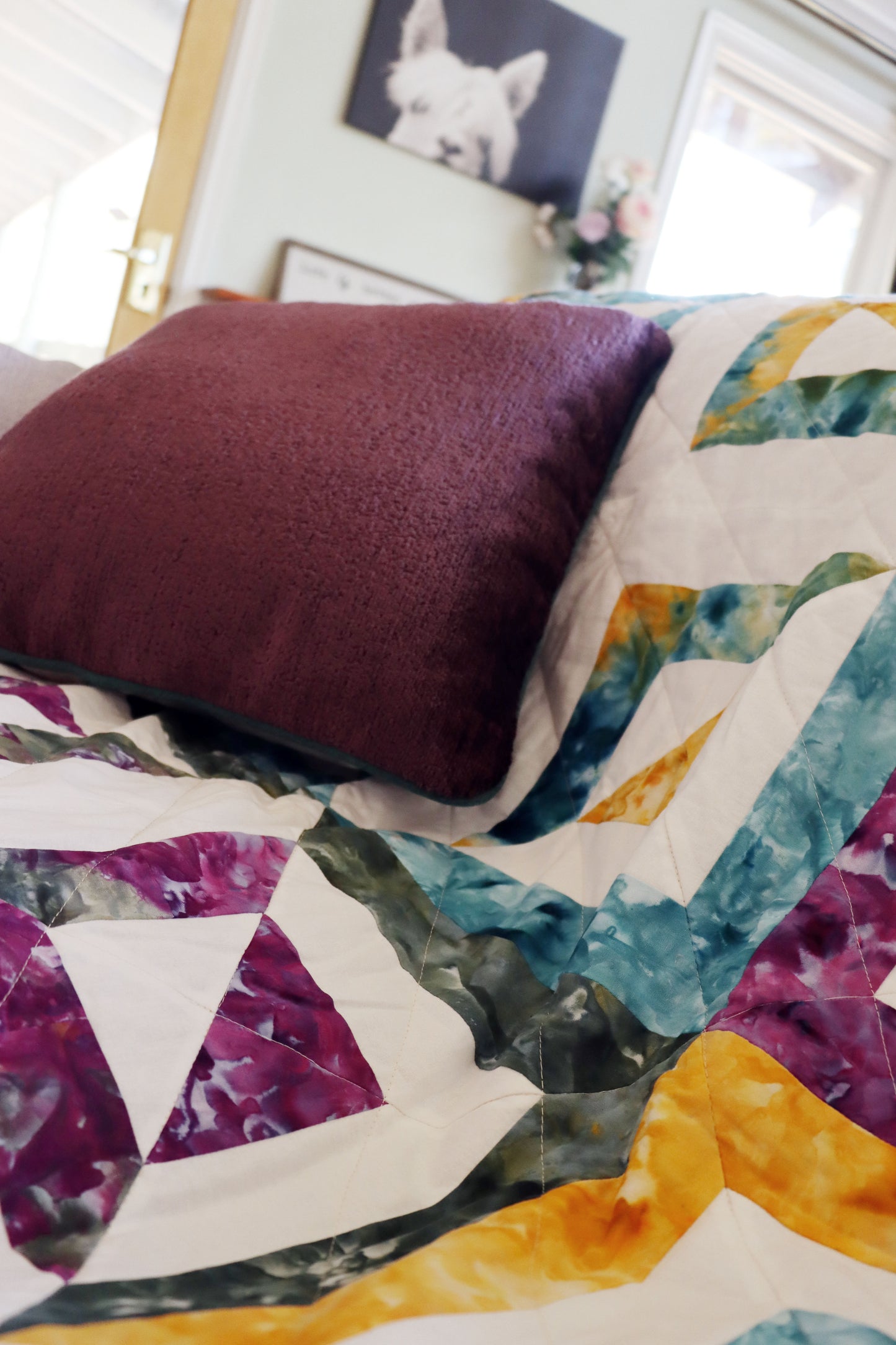 Colourful quilt with geometric patterns and a purple chenille cushion cover with contrasting piping on a sofa.
