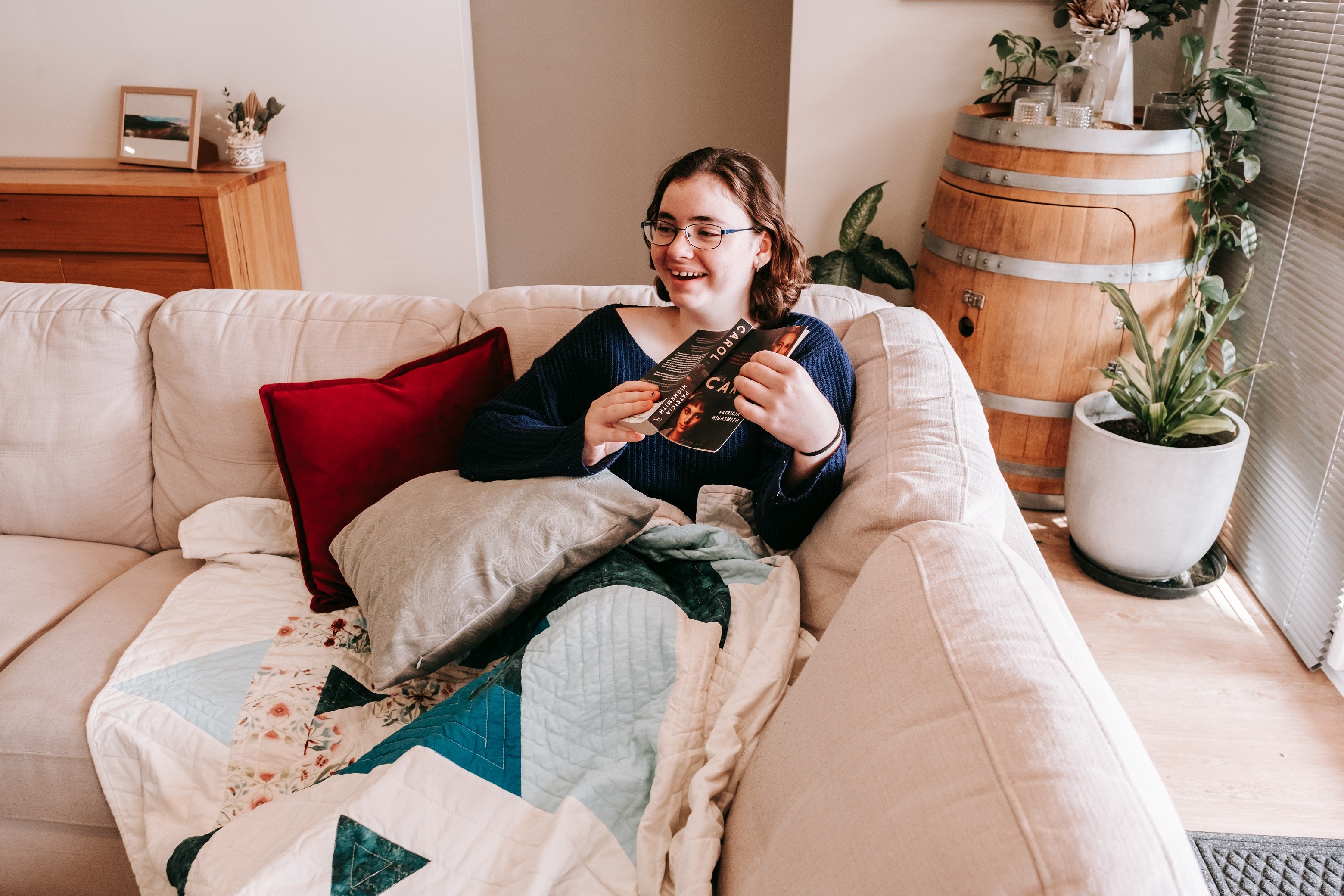 Lady sitting on the couch under a handmade quilt, holding a book and laughing 