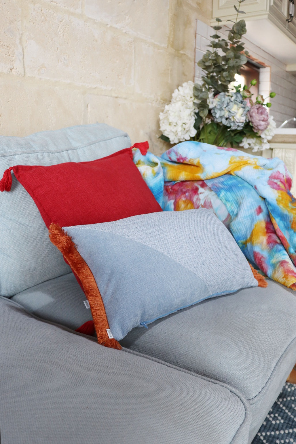 Gray sofa with an oblong cushion cover with cotton fringe and a floral arrangement on a stone wall background
