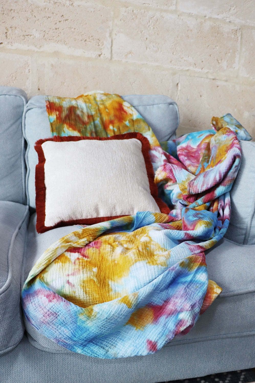 Colourful ice dye blanket draped over a gray sofa with a chenille cushion cover with cotton fringe