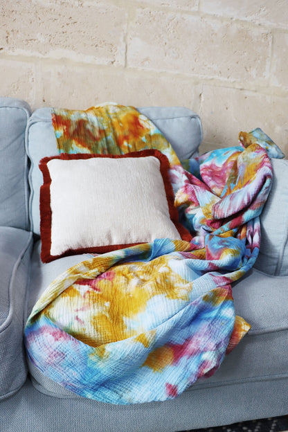 Colourful ice dye blanket draped over a gray sofa with a chenille cushion cover with cotton fringe