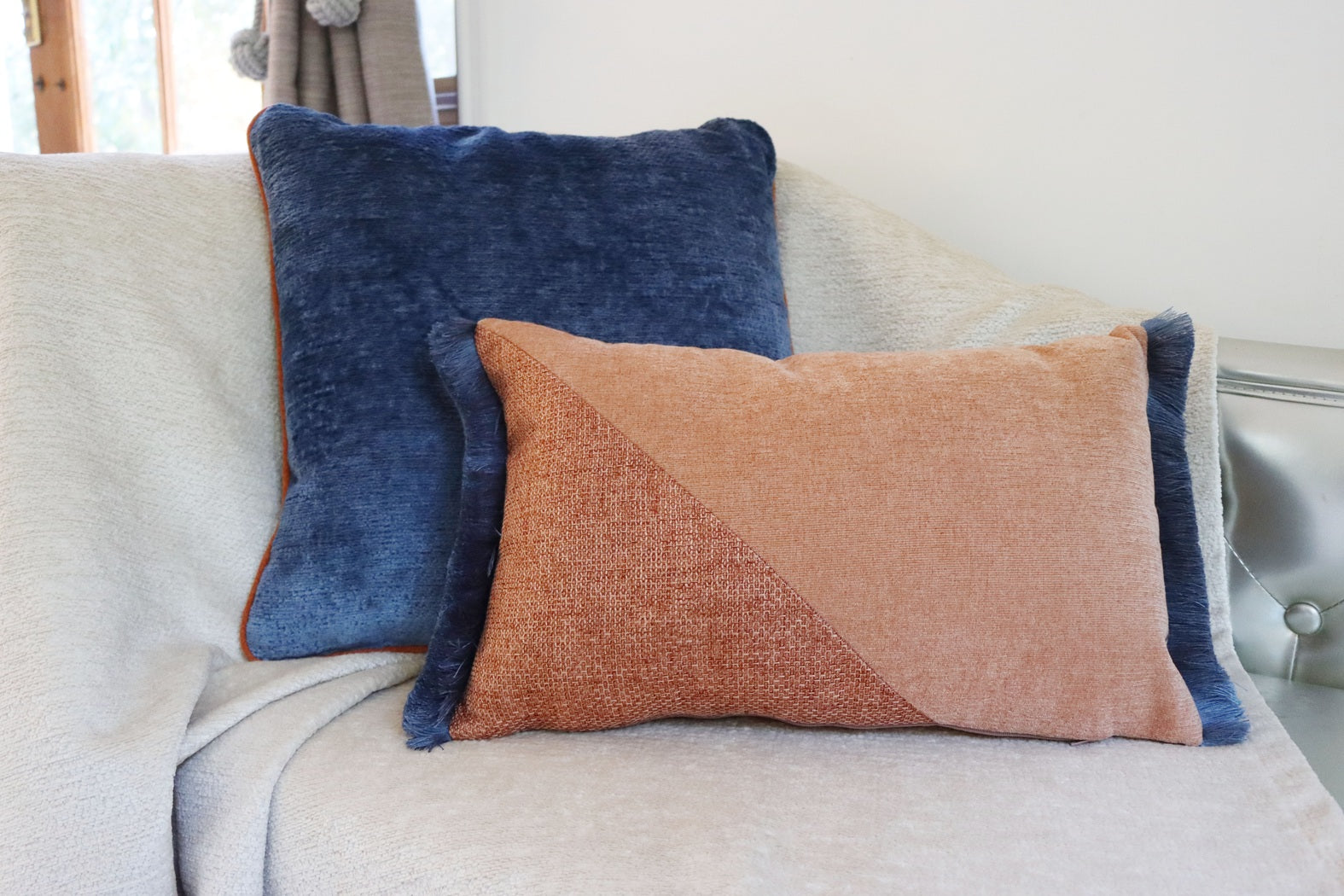 Two decorative pillows, one blue and one brown oblong cushion cover with cotton fringe, on a light-colored sofa.