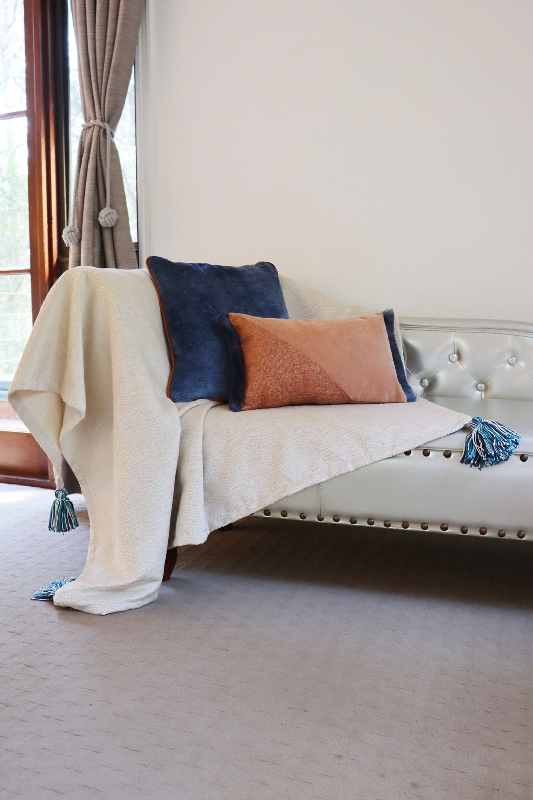 White sofa with decorative oblong cushion cover with cotton fringe and a blanket in a room with large windows.