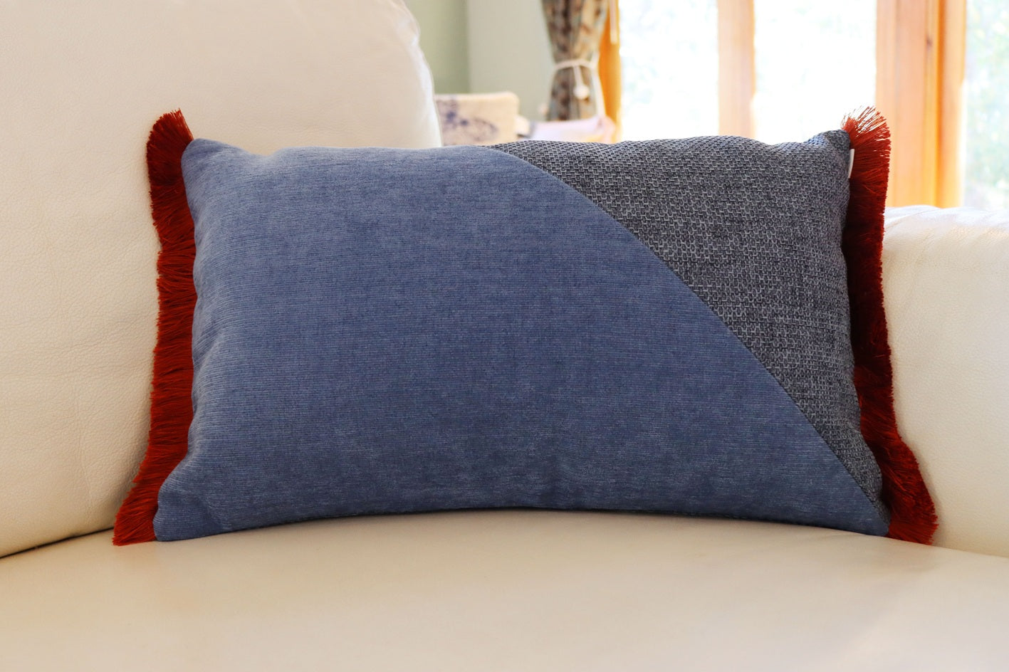 Blue and orange oblong cushion cover with cotton fringe on a beige sofa with a blurred indoor background