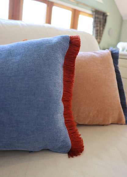 Blue and orange oblong cushion cover with cotton fringe on a couch.