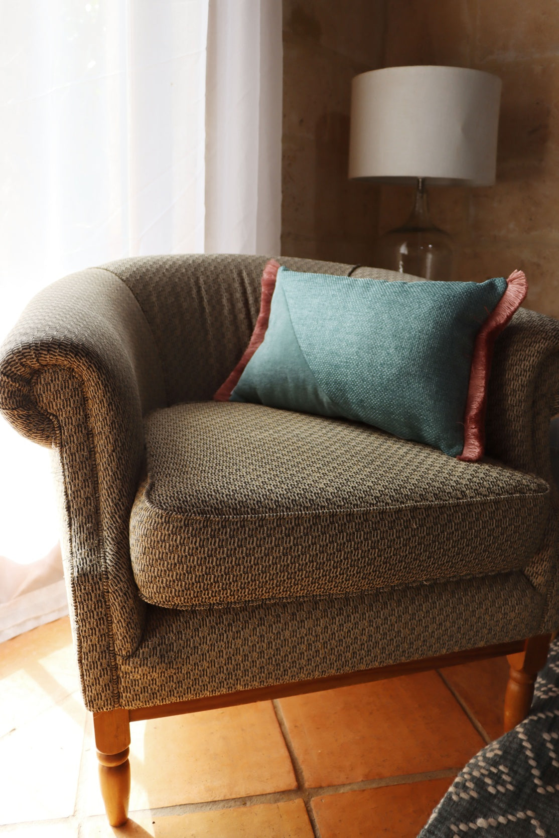 Brown armchair with a green oblong cushion cover with cotton fringe in a room with a lamp and window.