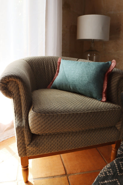 Brown armchair with a green oblong cushion cover with cotton fringe in a room with a lamp and window.
