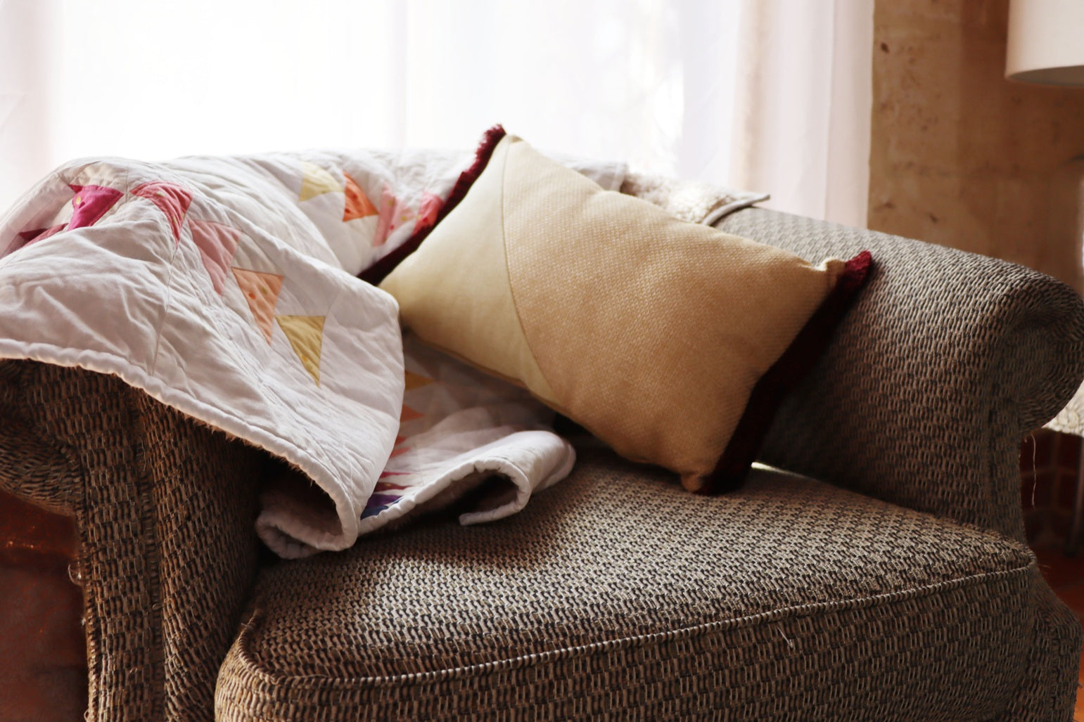 Brown armchair with a yellow oblong cushion cover with cotton fringe and colorful quilt in a softly lit room.