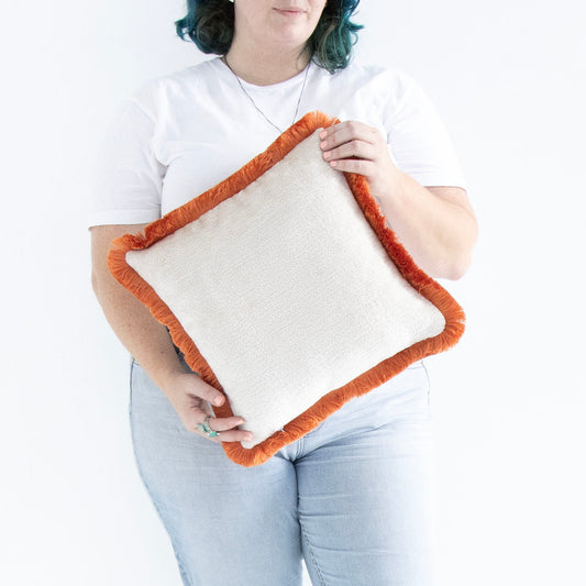 Person holding an orange chenille cushion cover with cotton fringe against a white background