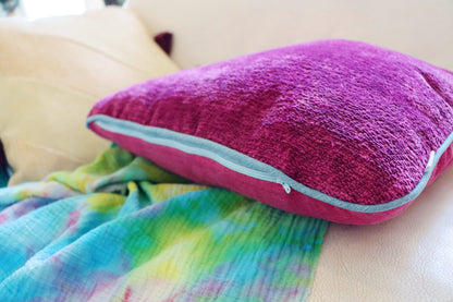Pink chenille cushion cover with contrasting piping with a colourful blanket on a beige surface