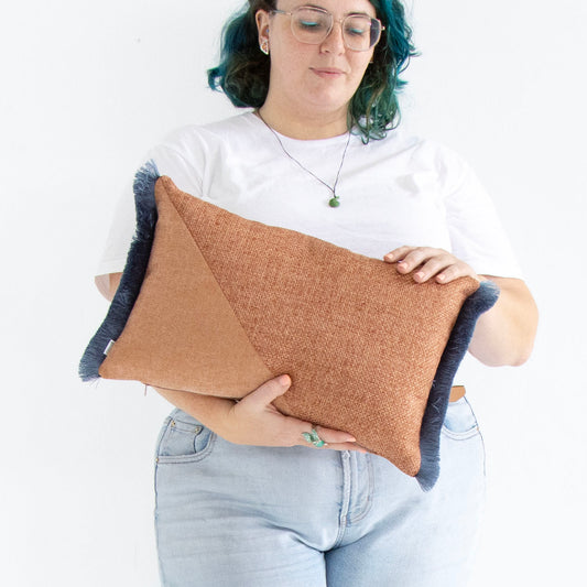 Person holding a brown and blue oblong cushion cover with cotton fringe against a white background
