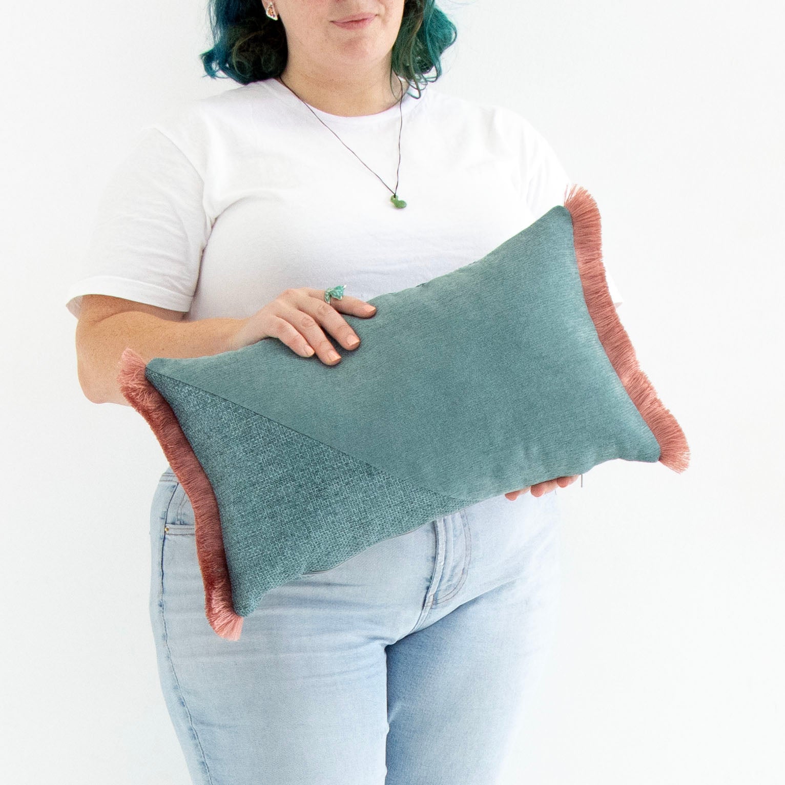 Person holding a teal oblong cushion cover with cotton fringe against a white background