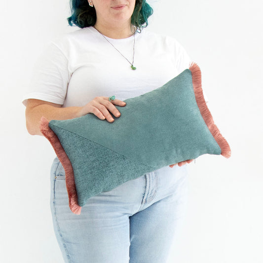 Person holding a teal oblong cushion cover with cotton fringe against a white background