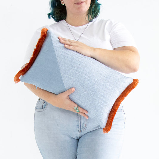 Person holding a blue and orange geometric oblong cushion cover with cotton fringe against a white background