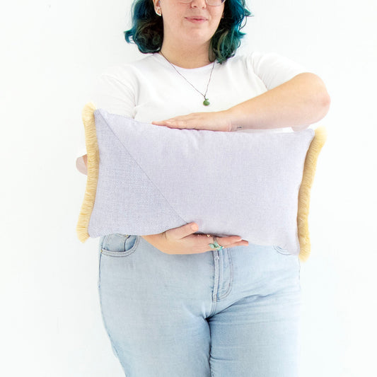 Person holding a light purple oblong cushion cover with cotton fringe against a white background