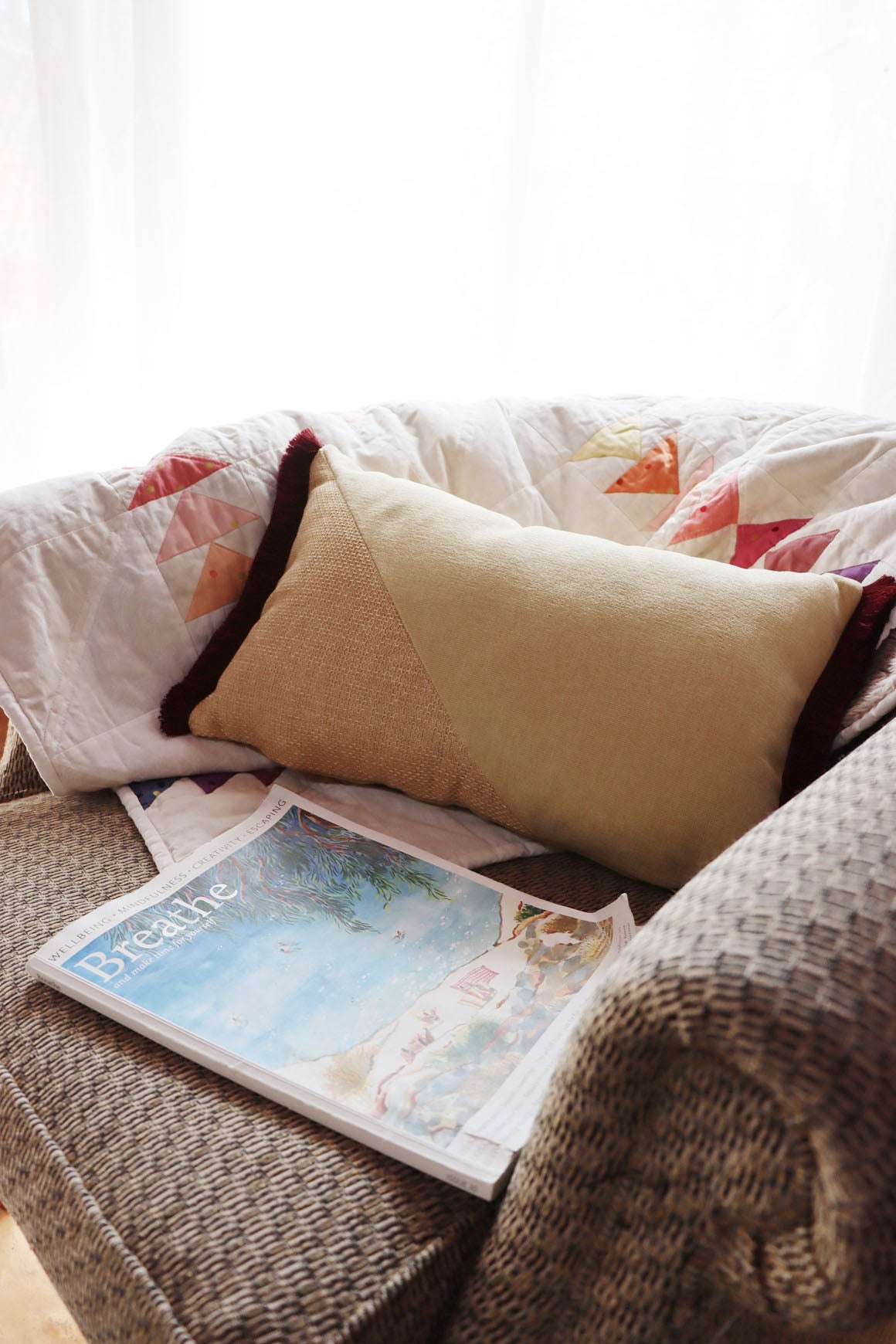 Cosy living room scene with a yellow and red oblong cushion cover with cotton fringe, and a handmade quilt, and magazine on a sofa.