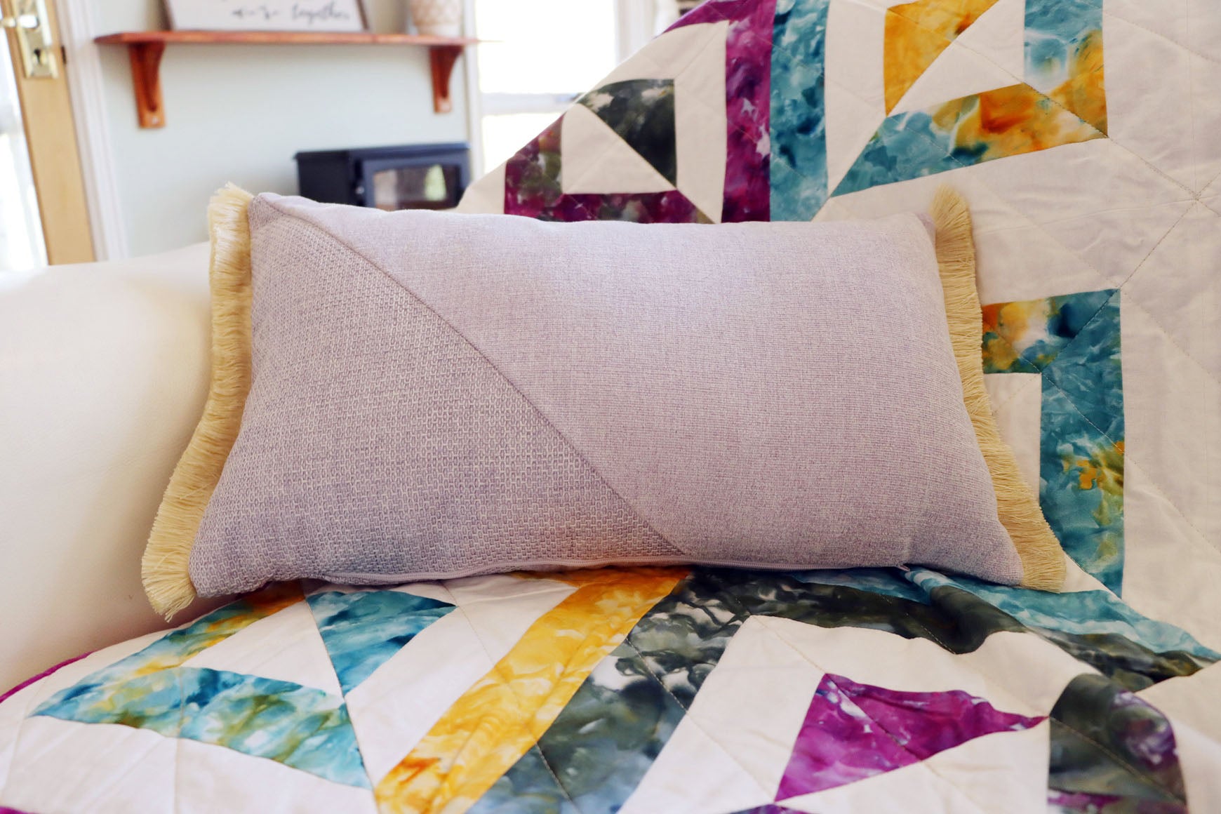 Purple and yellow oblong cushion cover with cotton fringe on a colorful quilted blanket