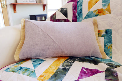 Purple and yellow oblong cushion cover with cotton fringe on a colorful quilted blanket